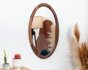May include: A brown oval-shaped wooden frame mirror with a beveled edge. The mirror is hanging on a white wall.