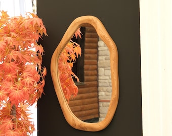 May include: An oval-shaped wooden mirror with a unique, organic frame. The mirror reflects a view of a wooden cabin and autumn foliage. The frame has a natural wood grain finish, adding a touch of rustic charm to the decor.