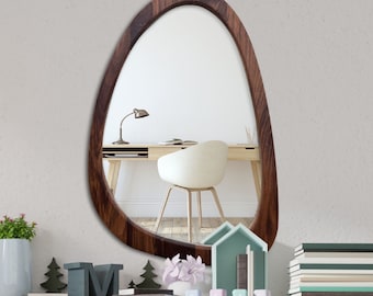 May include: A brown wooden oval mirror with a smooth, slightly irregular edge. The mirror is hanging on a white wall and reflects a desk with a chair and a lamp.