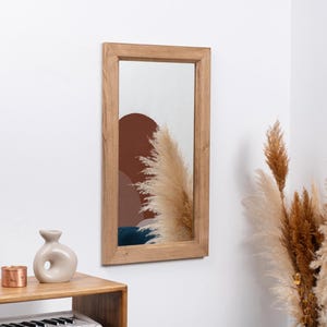 May include: A rectangular wooden framed mirror with a light brown wood frame. The mirror reflects a room with a brown and white abstract design and a white fluffy plant.