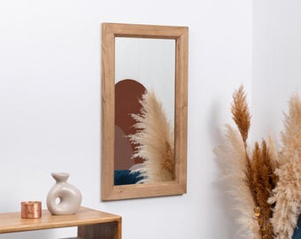 May include: A rectangular wooden framed mirror with a light brown wood frame. The mirror reflects a room with a brown and white abstract design and a white fluffy plant.