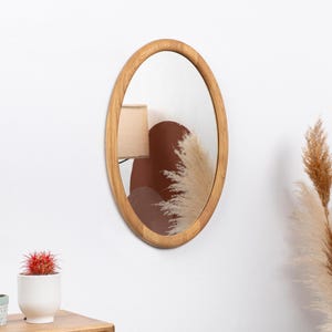 Può includere: Uno specchio rotondo con cornice in legno con finitura in legno naturale. Lo specchio è di forma ovale e ha un bordo bisellato. Lo specchio è appeso a una parete bianca.