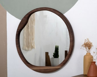 May include: A dark wood framed mirror with an irregular, organic shape. The mirror reflects a white wall and a potted plant.