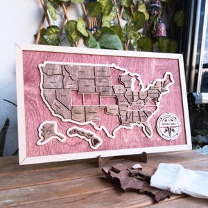 May include: A wooden map of the United States with each state outlined and labeled. The map is mounted on a pink wood background and has a compass with the text "Brown Family" in the bottom right corner.