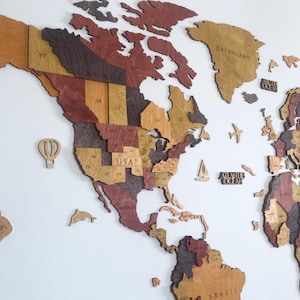 May include: A wooden world map featuring North and South America in shades of brown, red, and tan. Decorative wooden cutouts of a hot air balloon, airplane, sailboat, and the words "Atlantic Ocean" are included. The map is mounted on a white wall.