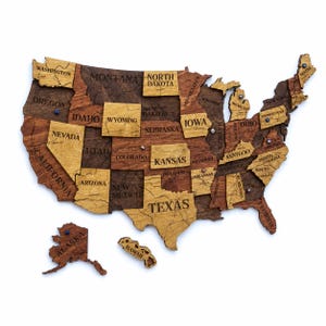 May include: A wooden wall map of the United States featuring individual state pieces in varying shades of brown. Each state is laser-engraved with its name and borders. The map includes Alaska and Hawaii as separate pieces with small metal pins.