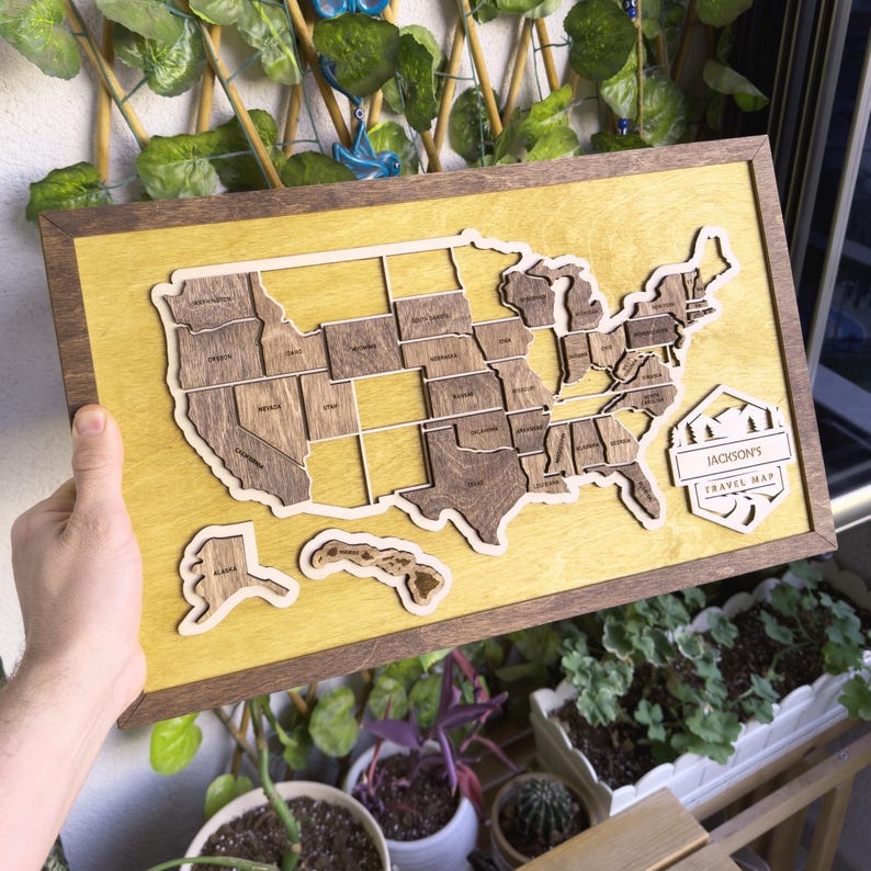 May include: A wooden map of the United States with each state outlined in a light brown color. The map is mounted in a brown frame with a yellow background. The text "Jackson's Travel Map" is printed on a brown octagon shape in the bottom right corner of the map.