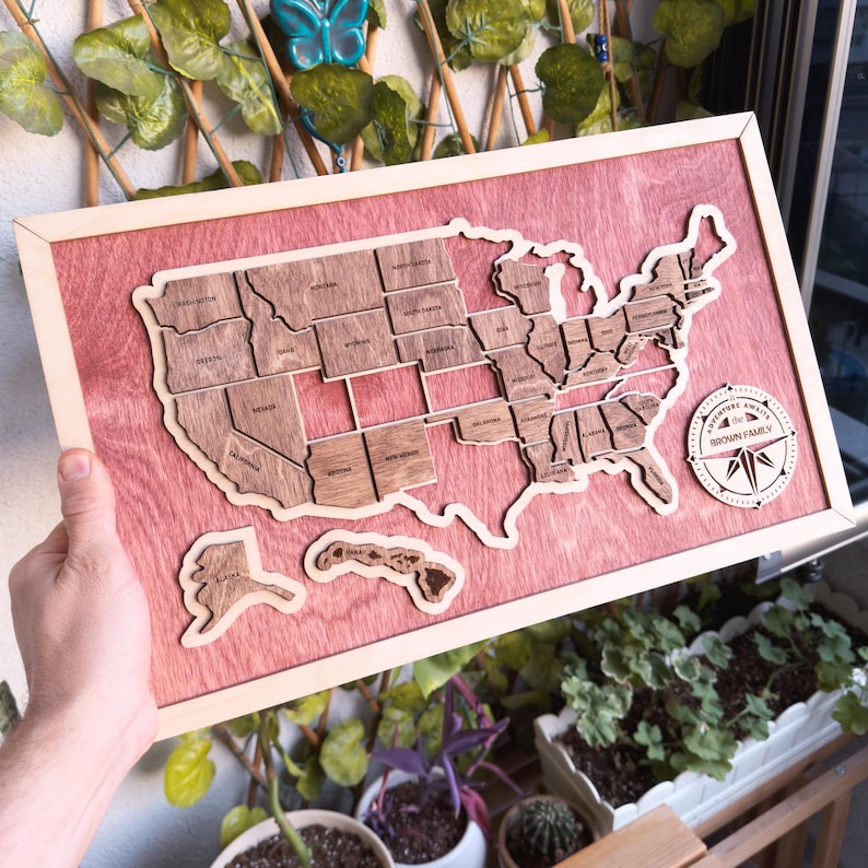 May include: A wooden map of the United States with each state outlined and labeled. The map is mounted on a red wood background with a compass in the lower right corner. The compass has the words "Explore Away" and "Brown Family" on it.
