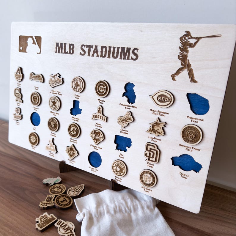May include: A wooden board with the text "MLB STADIUMS" and a baseball player silhouette. The board has cutouts for each MLB team's stadium, with the team's logo and stadium name printed below each cutout. The board is accompanied by a set of wooden team logo tokens.