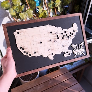 May include: A wooden map of the United States with baseballs marking each state. The map is framed in dark wood and has a baseball-themed logo that says "Baseball Ballpark Tour".