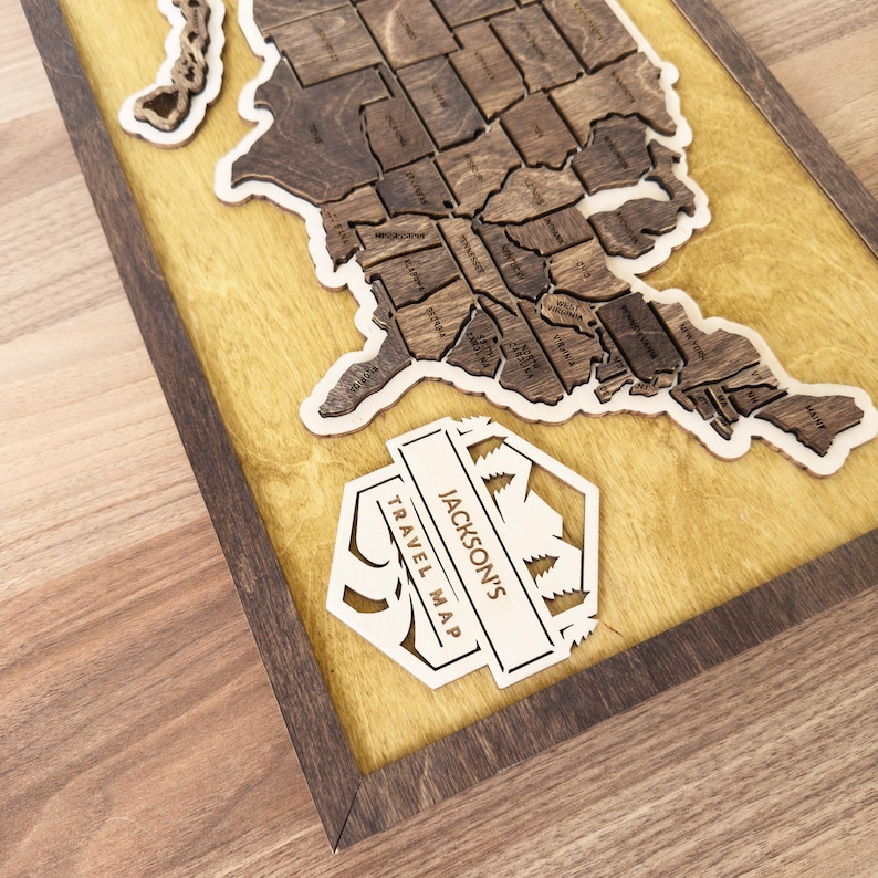 May include: A wooden map of the United States with a yellow background and a wooden frame. The map is divided into individual states and has a small wooden plaque with the text "JACKSON'S TRAVEL MAP" at the bottom right corner.