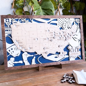 May include: A wooden map of the United States with baseball stadiums marked. The map is framed in dark wood and has a blue background. The map is on a stand and there are baseballs and a glove on the map. The text "Williams Family Baseball Stadium Bucket List" is on the map.