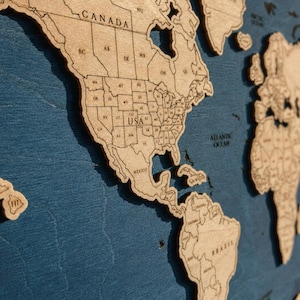 May include: A laser-cut wooden world map featuring light brown continents with engraved borders and labels for countries and states, including Canada, USA, and Mexico, mounted against a dark blue textured background with visible wood grain.