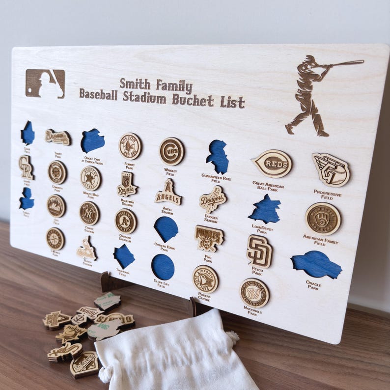 May include: A wooden board with the text "Smith Family Baseball Stadium Bucket List" and a baseball player silhouette. The board has a grid of 36 circles, each with a different baseball team logo. The logos are in various colors, including red, blue, white, and black. The board is on a table with a white cloth bag and a few loose logos.