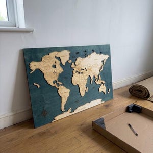 May include: A rectangular wooden world map wall art piece featuring light-colored laser-cut continents mounted onto a dark teal-stained wooden board. The map includes engraved country names and borders, with a compass rose in the bottom left corner.