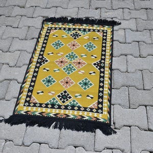 May include: A rectangular woven rug featuring a mustard yellow background with a repeating pattern of small diamonds in green, black, and light pink. The rug has a black border with white geometric accents and black fringe on both ends.