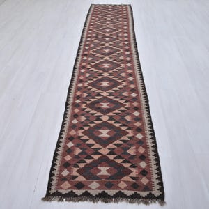 May include: A long, rectangular runner rug with a geometric pattern in shades of brown, beige, and black. The rug features diamond and triangle shapes, with a dark border and fringe at the ends. The rug is placed on a light-coloured wooden floor.