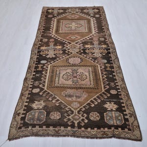 Puede incluir: Una alfombra rectangular de estilo vintage con un patrón geométrico central en tonos marrones, beige y tostados. La alfombra presenta un diseño simétrico con motivos repetidos y un borde marrón oscuro. La alfombra está colocada sobre una superficie blanca.