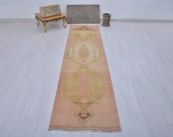 Puede incluir: Una alfombra larga y estrecha con fondo rosa y un patrón geométrico en tonos beige y verde. La alfombra tiene un borde con flecos.