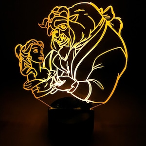 Può includere: Una luce notturna stampata in 3D di colore giallo che rappresenta Belle e la Bestia del film Disney La Bella e la Bestia. La luce è accesa e illumina le figure.