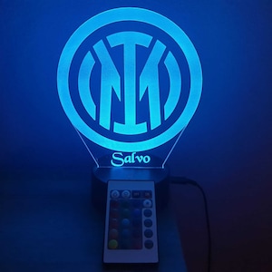 Può includere: Una luce notturna stampata in 3D blu e bianca con il logo dell'Inter e il nome "Salvo" sotto. La luce è su una base nera con un telecomando.