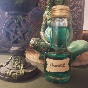 May include: A small green glass bottle with a gold lid and a label that says "chance". The bottle is sitting on a wooden surface in front of a green statue.