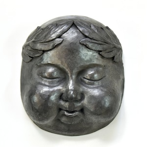 Könnte beinhalten: Eine graue Keramikskulptur eines Gesichts mit geschlossenen Augen und einem Kranz aus Blättern auf dem Kopf.