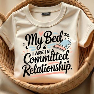 Puede incluir: Una camiseta de color crema con un estampado gráfico que dice "My Bed & I are in a committed relationship." El gráfico incluye una cama con una almohada y un corazón.