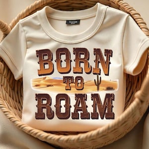 May include: Cream-colored t-shirt with the words "BORN TO ROAM" in brown, Western-style lettering. The design includes a desert landscape with a cactus. The shirt is displayed in a woven basket.