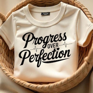 May include: A cream-colored t-shirt with short sleeves and a black graphic that reads "Progress over Perfection" with a heartbeat symbol.
