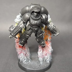 May include: A dark grey miniature figurine of a futuristic armoured soldier in flight, equipped with a jetpack and two handheld blasters. The figure is suspended above a circular base by sculpted smoke effects painted in shades of red, orange, and white.
