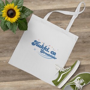 Peut inclure: Sac fourre-tout blanc avec le texte "Kukki. co Streetwear" en bleu et une paire de baskets vertes. Un tournesol jaune avec des feuilles vertes est dans le coin supérieur gauche. Le sac a deux anses blanches.