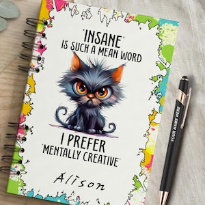 以下が含まれることがあります： カラフルなペイントスプラッターの縁取り、猫のイラスト、'INSANE' IS SUCH A MEAN WORD I PREFER 'MENTALLY CREATIVE'というテキストとAlisonという名前が記された白い表紙のリングノート。黒とローズゴールドのペンが添えられています。