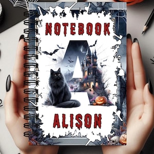 Puede incluir: Un cuaderno negro y blanco con un tema de Halloween. La portada presenta un gato negro, un castillo espeluznante y la letra "A" con el nombre "Alison" escrito debajo. El cuaderno tiene una encuadernación en espiral.