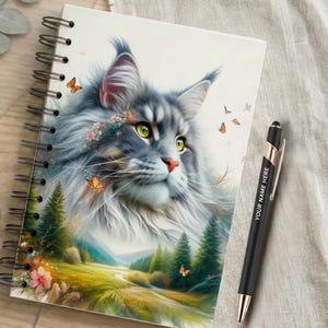 May include: A spiral-bound notebook featuring a detailed illustration of a fluffy grey cat with green eyes, surrounded by small orange butterflies, set against a landscape of green pine trees and a meadow. A black pen with the text YOUR NAME HERE is included.