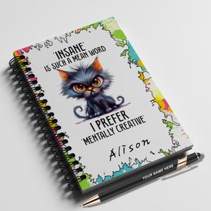Puede incluir: Un cuaderno de espiral con una ilustración de gato de dibujos animados y el texto "INSANE IS SUCH A MEAN WORD I PREFER 'MENTALLY CREATIVE' Alison". Un bolígrafo negro con detalles dorados está al lado del cuaderno, con el texto "YOUR NAME HERE".