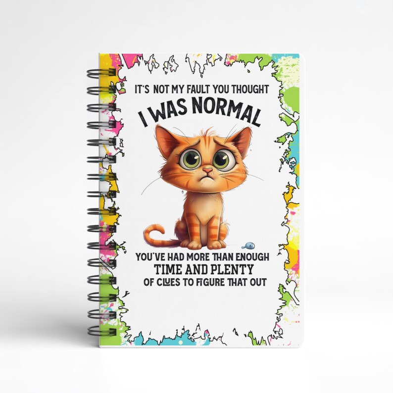 K&ouml;nnte beinhalten: Spiralgebundenes Notizbuch mit farbenfrohem Rand und dem Text "IT'S NOT MY FAULT YOU THOUGHT I WAS NORMAL." Das Cover zeigt eine Cartoon-Katze in Orange mit gro&szlig;en gr&uuml;nen Augen. Der Text "YOU'VE HAD MORE THAN ENOUGH TIME AND PLENTY OF CLUES TO FIGURE THAT OUT" ist ebenfalls vorhanden.