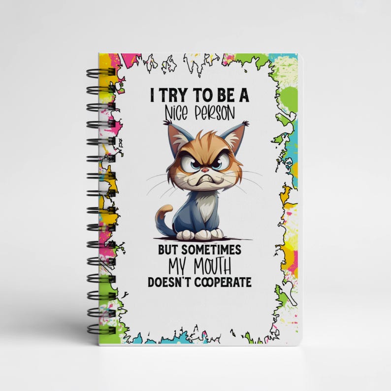 K&ouml;nnte beinhalten: Ein spiralgebundenes Notizbuch mit wei&szlig;em Einband. Es zeigt eine Cartoon-Katze mit w&uuml;tendem Gesichtsausdruck und dem Text: "I TRY TO BE A NICE PERSON BUT SOMETIMES MY MOUTH DOESN'T COOPERATE." Die R&auml;nder sind mit bunten Farbspritzern verziert.