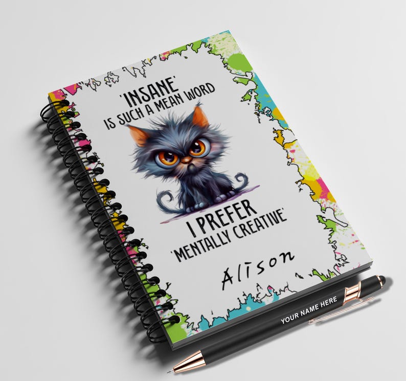 K&ouml;nnte beinhalten: Ein Notizbuch mit einer Cartoon-Katzen-Illustration und dem Text "INSANE IS SUCH A MEAN WORD I PREFER 'MENTALLY CREATIVE' Alison". Ein schwarzer Stift mit goldenen Akzenten liegt neben dem Notizbuch, mit dem Text "YOUR NAME HERE".