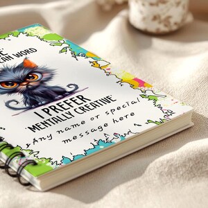 K&ouml;nnte beinhalten: Ein Spiralnotizbuch mit einem farbenfrohen Einband, der eine Cartoon-Katze mit gro&szlig;en orangefarbenen Augen zeigt. Der Text auf dem Einband lautet "I PREFER MENTALLY CREATIVE" und "Any name or special message here". Die Seitenr&auml;nder sind mit bunten Designs verziert.