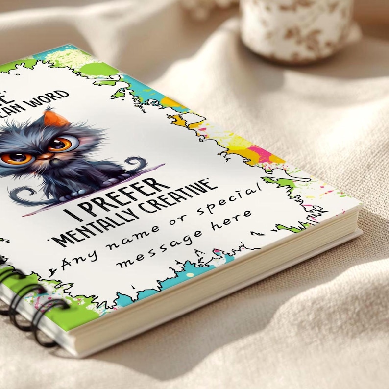 K&ouml;nnte beinhalten: Ein Spiralnotizbuch mit einem farbenfrohen Einband, der eine Cartoon-Katze mit gro&szlig;en orangefarbenen Augen zeigt. Der Text auf dem Einband lautet "I PREFER MENTALLY CREATIVE" und "Any name or special message here". Die Seitenr&auml;nder sind mit bunten Designs verziert.