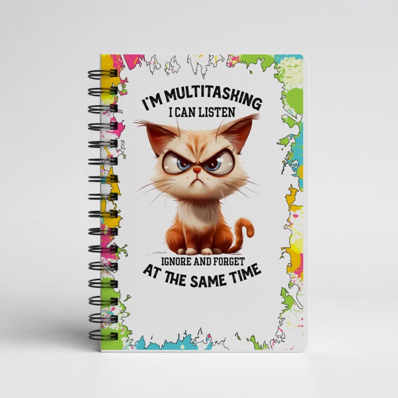 K&ouml;nnte beinhalten: Ein spiralgebundenes Notizbuch mit einer Cartoon-Katzen-Illustration. Die Katze mit Brille hat einen w&uuml;tenden Gesichtsausdruck. Der Text auf dem Cover lautet "I'M MULTITASKING I CAN LISTEN IGNORE AND FORGET AT THE SAME TIME". Das Notizbuch hat einen farbenfrohen Rand.