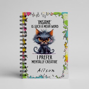 K&ouml;nnte beinhalten: Ein Spiralnotizbuch mit einer Cartoon-Katzenillustration. Die Katze ist grau mit orangefarbenen Ohren und gro&szlig;en gelben Augen. Der Text auf dem Cover lautet: "'INSANE' IS SUCH A MEAN WORD, I PREFER 'MENTALLY CREATIVE' Alison."