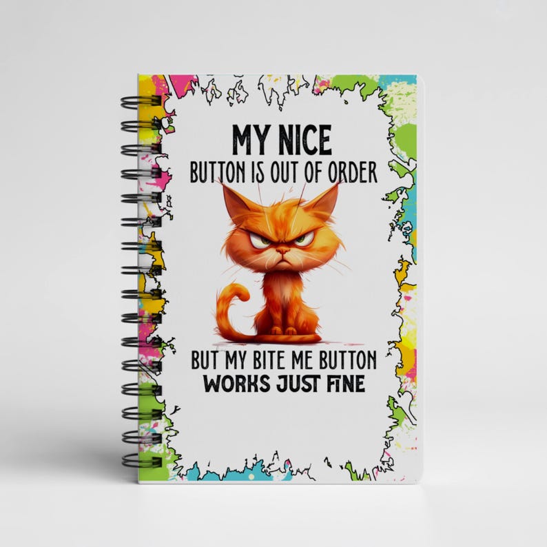 K&ouml;nnte beinhalten: Ein spiralgebundenes Notizbuch mit wei&szlig;em Einband, das eine Illustration einer w&uuml;tenden orangefarbenen Katze zeigt. Der Text lautet: "MY NICE BUTTON IS OUT OF ORDER BUT MY BITE ME BUTTON WORKS JUST FINE." Das Notizbuch hat einen bunten, abstrakten Rand.