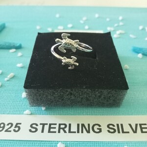 May include: A silver ring featuring two turtle designs. The ring is displayed on a black velvet background. The text "925 STERLING SILVER" is visible below the ring.