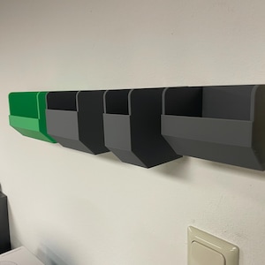 May include: Five wall-mounted storage bins, four are grey and one is green. The bins are made of plastic and have a rectangular shape. The bins are mounted on a white wall.