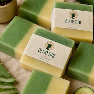 May include: Five rectangular bars of soap, each featuring a dual-tone design with one half in solid green and the other in pale yellow. Two bars are wrapped in a cream-coloured paper label printed with the text Aloe Bar Handcrafted Net Wt 4oz (113g).