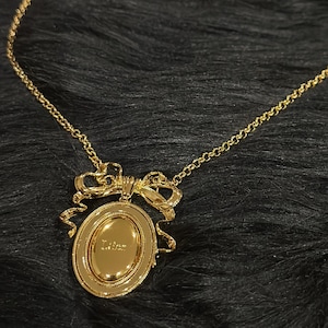 May include: A gold-toned necklace featuring an oval pendant with a central embossed design and a decorative bow detail at the top. The pendant is suspended from a delicate gold-toned chain with a lobster clasp closure at the back of the neck.