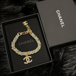 Könnte beinhalten: Ein goldfarbenes Kettenarmband, das mit hellblauem Jeansstoff durchflochten ist, mit einem hängenden CC-Logo-Anhänger, der mit klaren Kristallen besetzt ist, und einem kleineren CC-Logo am Verschluss. Das Armband liegt in einer schwarzen Box mit Chanel-Logo.