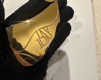 May include: A gold-colored pendant with the letters "DV" in a stylized font. The pendant is held by a gloved hand, and the background includes a "CHANEL" book.